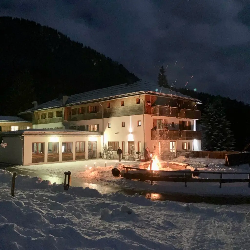 Verschneite Landschaft bei Nacht mit einem großen, beleuchteten Gebäude im Hintergrund. Im Vordergrund brennt ein Lagerfeuer, umgeben von Bänken. Der Himmel ist dunkel, und um das Feuer steigen Funken auf.