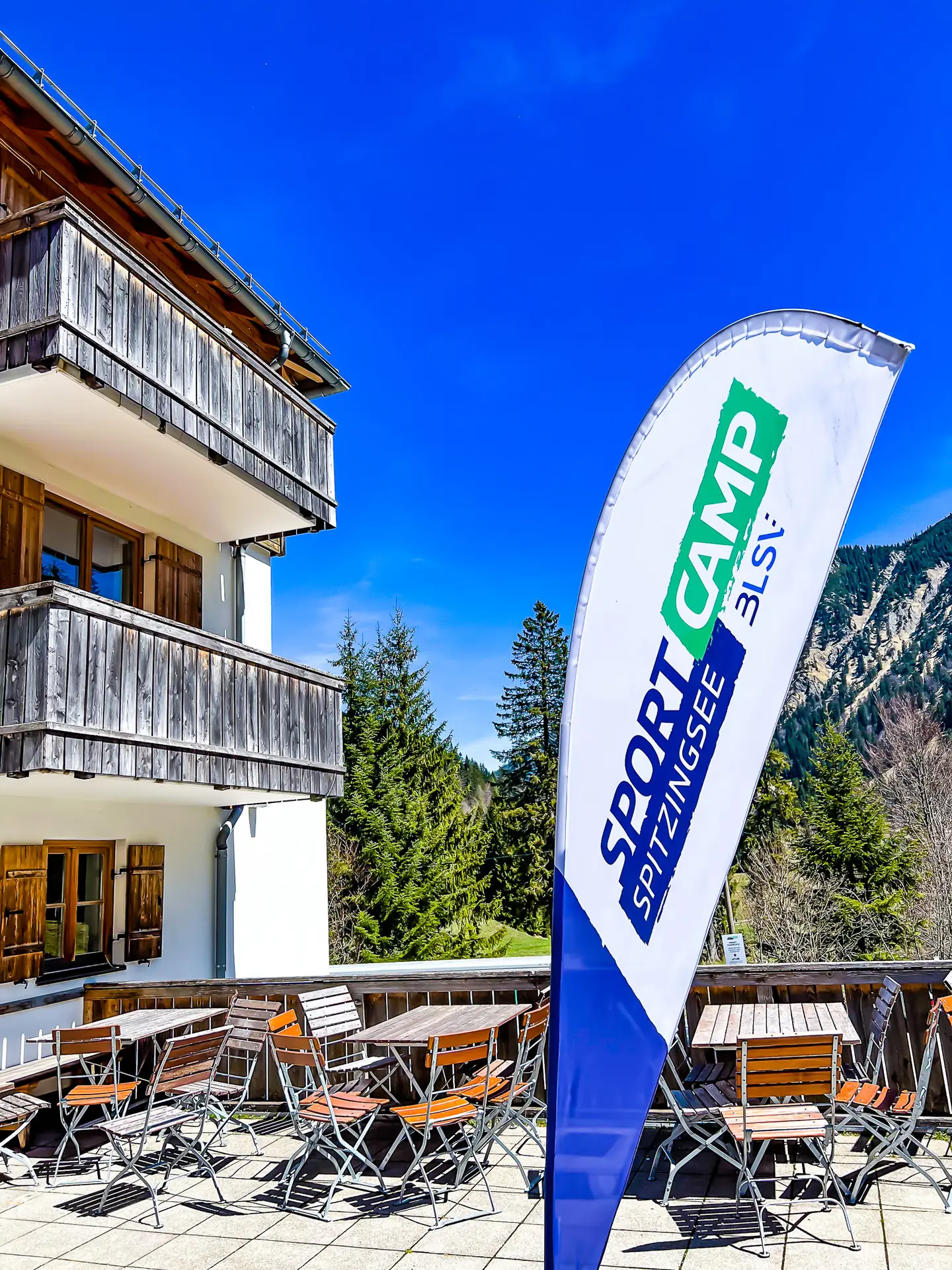 IMG_4642 Außenbereich eines Gebäudes im alpinen Stil mit Holzbalkon und Fensterläden. Davor befinden sich leere Tische und Stühle auf einer Terrasse. Eine Fahne mit der Aufschrift "Sportcamp Spitzingsee BLSV" weht im Vordergrund. Im Hintergrund sind grüne Bäume und ein blauer Himmel sichtbar.