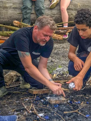 08be2384-7c91-463e-b35e-dbdda740f10f Ein Mann und ein Junge hocken auf dem Boden und entzünden gemeinsam ein Lagerfeuer. Im Hintergrund sind weitere Personen sichtbar, die auf einem umgestürzten Baumstamm sitzen. Um sie herum liegen Stöcke und Holzteile auf dem Boden. Der Mann zeigt dem Jungen, wie man das Feuer entzündet.