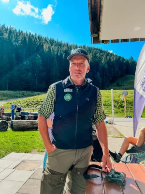 IMG_0675 Mann in Ranger-Kleidung steht vor einer Waldkulisse unter blauem Himmel. Er trägt eine Mütze und hat ein Fernglas auf einem Holztisch vor sich liegen. Im Hintergrund sind mehrere Menschen sowie Holzbänke zu sehen.
