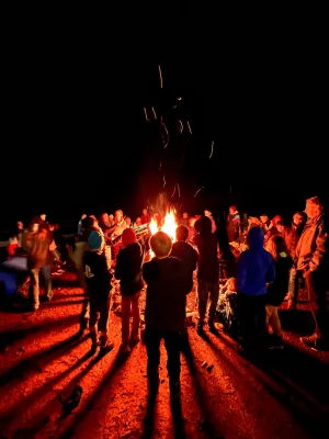 IMG_1908 Eine Gruppe von Menschen, darunter Erwachsene und Kinder, stehen in der Dunkelheit um ein loderndes Lagerfeuer. Funken steigen in den Nachthimmel, und das Feuer wirft lange Schatten auf den erdigen Boden. Die Szene wirkt warm und gesellig.