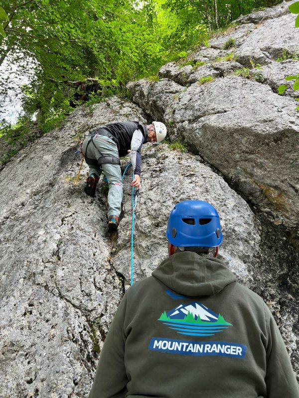 IMG_7445 Eine Person klettert mit einem Seil gesichert an einem felsigen Hang. Sie trägt einen Kletterhelm. Eine zweite Person, ebenfalls mit Helm, steht unten und schaut zu. Auf dem Rücken ihrer Jacke steht "Mountain Ranger". Die Umgebung ist von grünem Laub umgeben.