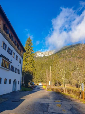 WhatsApp Image 2024-11-18 at 10.04.19 (1) Straße mit Schildern und Absperrungen in einer ländlichen Gegend, flankiert von einem großen Gebäude mit blauen Fensterläden links, dichten Wäldern und schneebedeckten Bergen im Hintergrund, bei klarem, blauem Himmel.