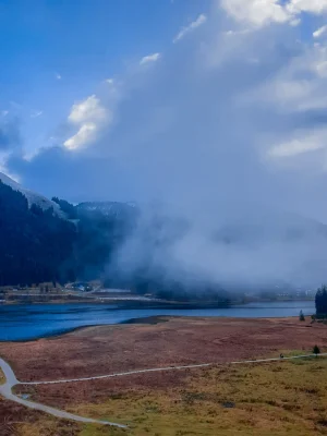 WhatsApp Image 2024-11-18 at 10.04.19 Wolkenverhangener Himmel über einem ruhigen See in einer Berglandschaft. Der Vordergrund zeigt eine weite Wiese mit kleinen Wegen, während im Hintergrund bewaldete Hügel und Nebel zu sehen sind.
